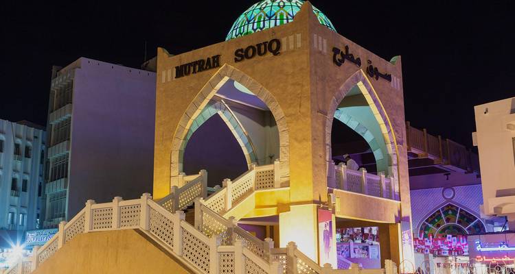 Vista nocturna de la entrada de arco iluminada del Souq de Mutrah con su cúpula de azulejos turquesas y su escalera ornamentada.