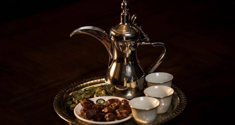 Cafetera tradicional omaní con tazas pequeñas, dátiles y cardamomo dispuestos en una bandeja de latón sobre un fondo oscuro.