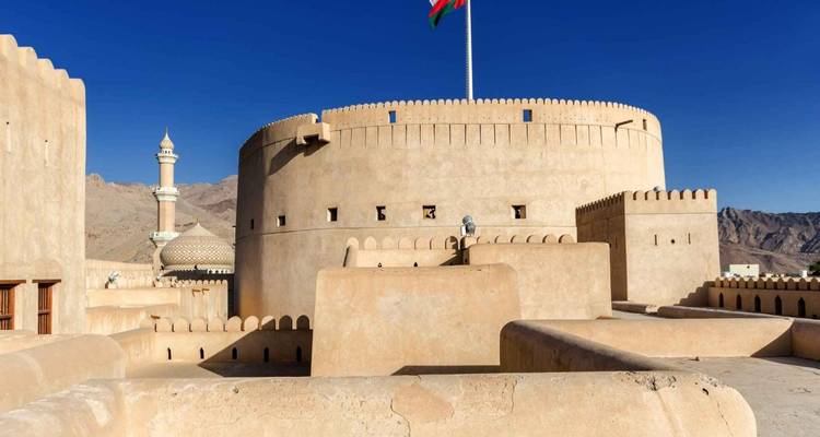 Torre circular iluminada por el sol del Fuerte de Nizwa ondeando la bandera omaní, con montañas desérticas y un minarete de mezquita de fondo.