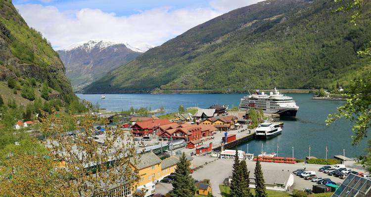 Village de fjord avec un navire de croisière amarré à côté de bâtiments aux toits rouges et de pics enneigés au loin.