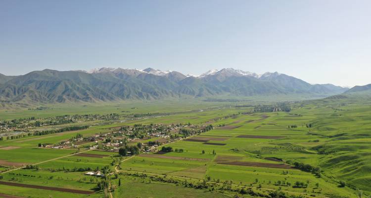 Wide aerial view of green valley fields backed by snow-dusted Kyrgyz mountains.