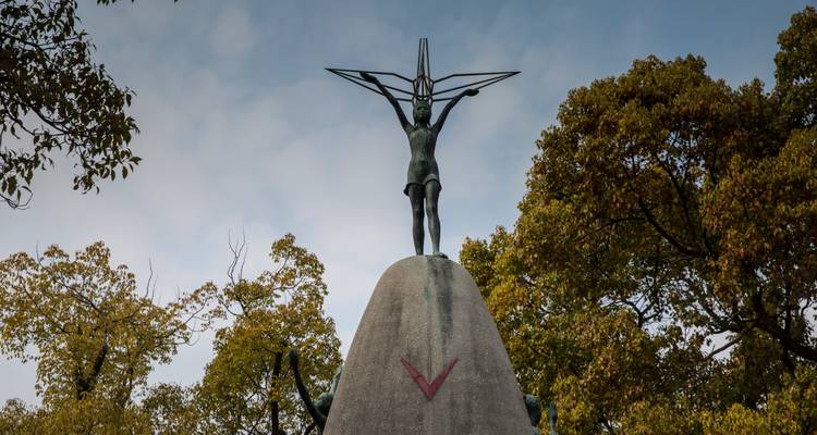 Monument de la Paix pour les Enfants, statue d'une fillette soulevant une grue en papier, encadrée par des arbres verts et un ciel couvert.