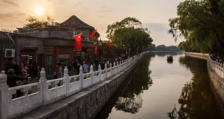 Historic Beijing hutong canal at sunset with Chinese flags and people strolling along the water.
