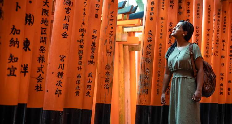 Traveler gazes upward while standing among rows of vivid orange torii gates with Japanese calligraphy.
