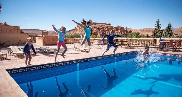 Niños y adultos saltando en el aire mientras se lanzan a una piscina azul brillante junto a un hotel kasbah en el desierto.