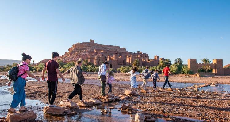 Viajeros cruzando piedras de salto a través de un río poco profundo con la fortaleza de adobe de Ait Benhaddou elevándose detrás.