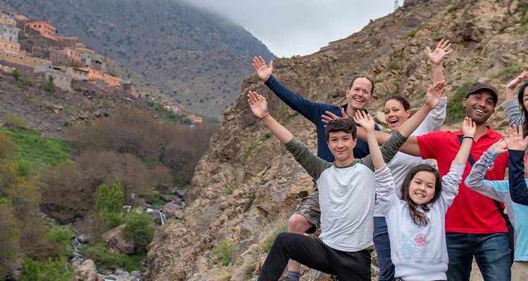 La familia saludando con entusiasmo en una cornisa rocosa de montaña con un pueblo bereber y picos brumosos detrás.