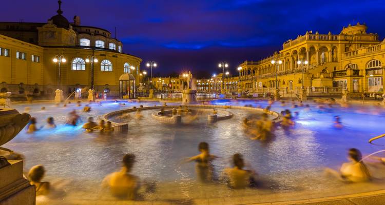 Photographie à longue exposition de nuit des Bains thermaux Széchenyi montrant des personnes profitant des bassins fumants sous le crépuscule bleu.