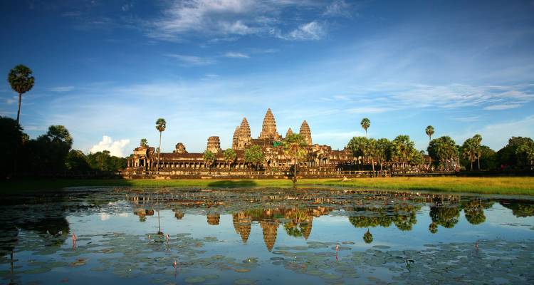 La silhouette iconique d'Angkor Wat se reflétant dans un étang couvert de nénuphars au lever du soleil sur un ciel bleu éclatant.
