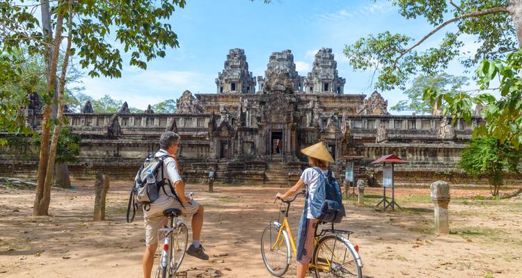 Deux cyclistes font une pause sous des arbres offrant de l'ombre face aux ruines d'un complexe de temples d'Angkor.