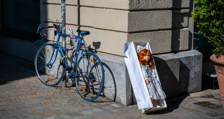 Street corner with blue bicycle and red-haired doll in white coffin leaning on wall.