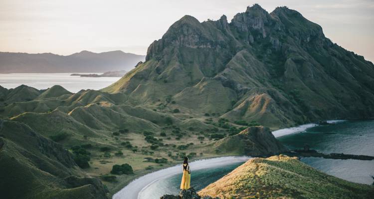 A lone woman in a yellow dress stands on a ridge overlooking the dramatic green ridges and turquoise bays of an island landscape.