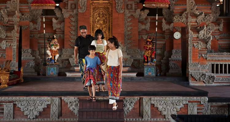 A family dressed in colorful sarongs descends the steps inside a richly carved Balinese temple hall.