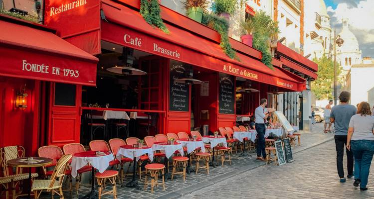 Café parisino típico con toldos rojos, sillas de ratán y clientes disfrutando de bebidas en una calle empedrada.