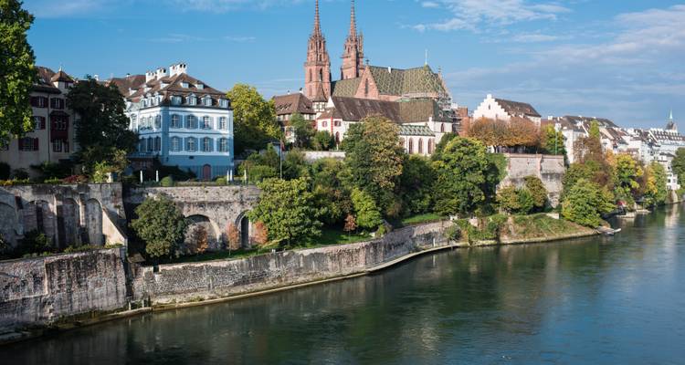 Malerische Flussufferansicht von Basel mit historischen Kathedraltürmen und farbenfrohen Gebäuden, die sich im Rhein unter einem strahlend blauen Himmel spiegeln.