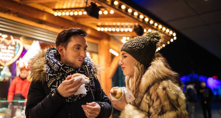Jong stel in winterkleding dat geniet van snacks bij een helder verlichte kersmarktstand