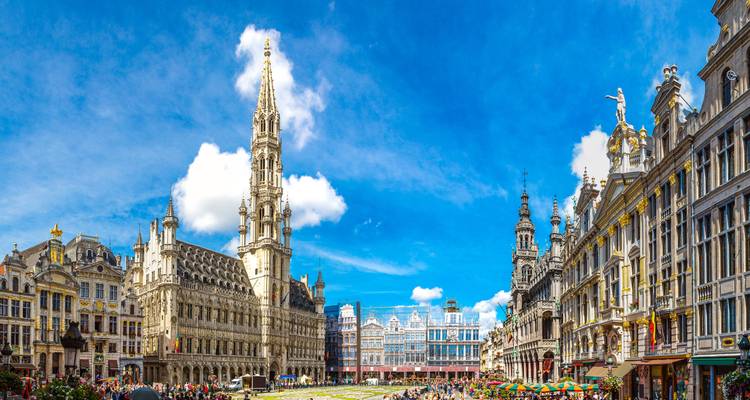 Panoramischer Überblick über den Brüsseler Großen Platz mit aufragendem gotischen Turm, prunkvollen Zunfthäusern und strahlend blauem Himmel.