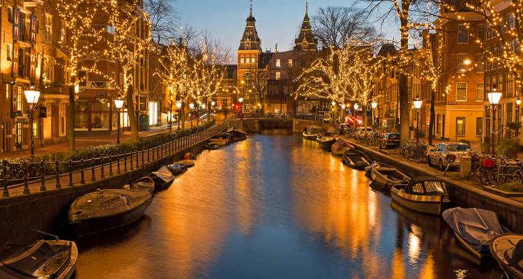 Canal de Ámsterdam al atardecer bordeado de lucecitas de hada titilantes y agua dorada reflejada.