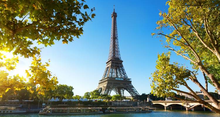 La Torre Eiffel elevándose sobre el Sena con barcos turísticos y árboles verdes iluminados por el sol.
