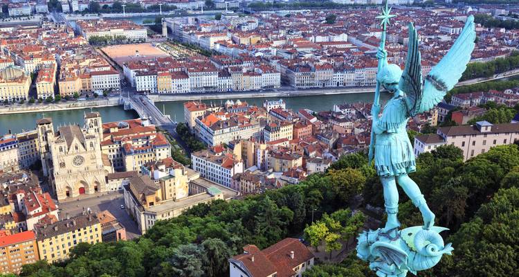 Estatua de arcángel de bronce que domina una extensa ciudad europea con dos ríos y tejados rojos.