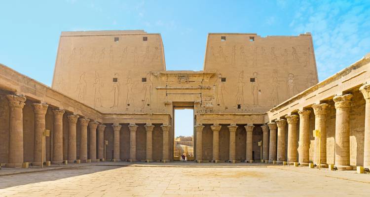 Amplio patio de un antiguo templo egipcio alineado con columnas ordenadas y pilonos cubiertos de jeroglíficos bajo un cielo brillante.