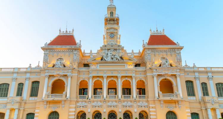 Vue de face diurne de l'Hôtel de Ville de Ho Chi Minh-Ville avec un ciel bleu dégagé mettant en évidence sa façade de couleur crème et ses toits rouges.