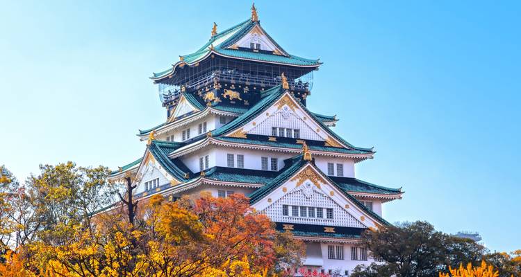 Osaka Castle towers above colorful autumn foliage against a bright blue sky.