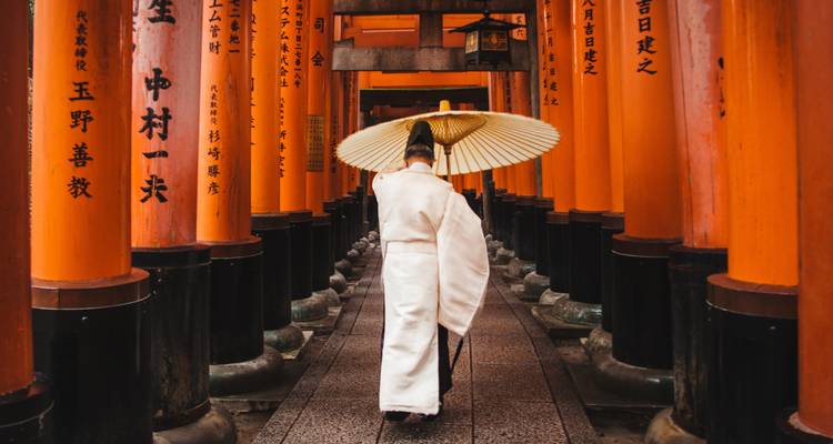 A Shinto priest in white robes walks through the vermilion torii gates of Fushimi Inari Shrine.