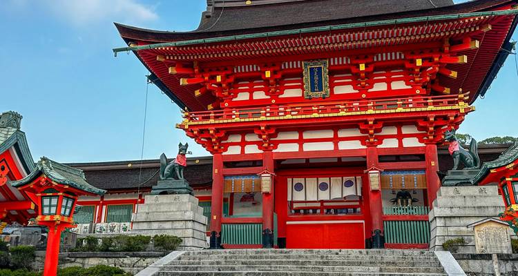Bright red main gate of a Shinto shrine with fox statues flanking the steps under a blue sky.