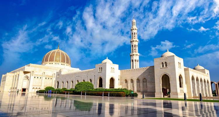 Gran Mezquita del Sultán Qaboos de mármol blanco con una cúpula ornamentada y un minarete bajo un vibrante cielo azul.