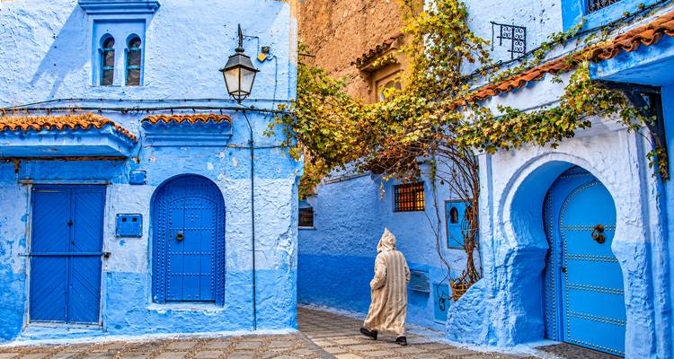 Una figura encapuchada pasa por puertas azules brillantemente pintadas y muros cubiertos de hiedra en Chefchaouen.