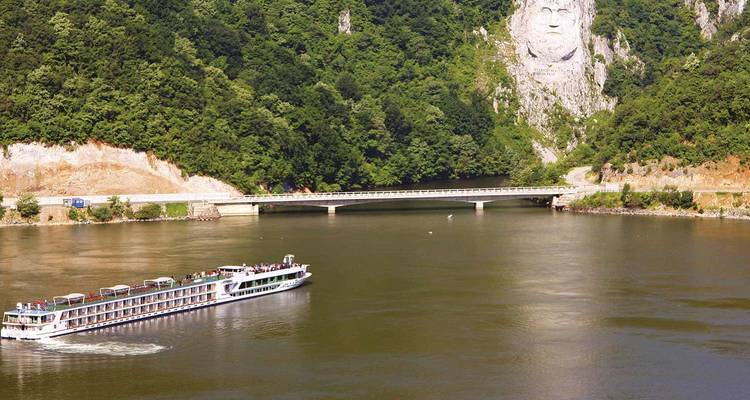 A river cruise ship glides past forested cliffs featuring the giant rock carving of King Decebalus at the Iron Gates gorge.