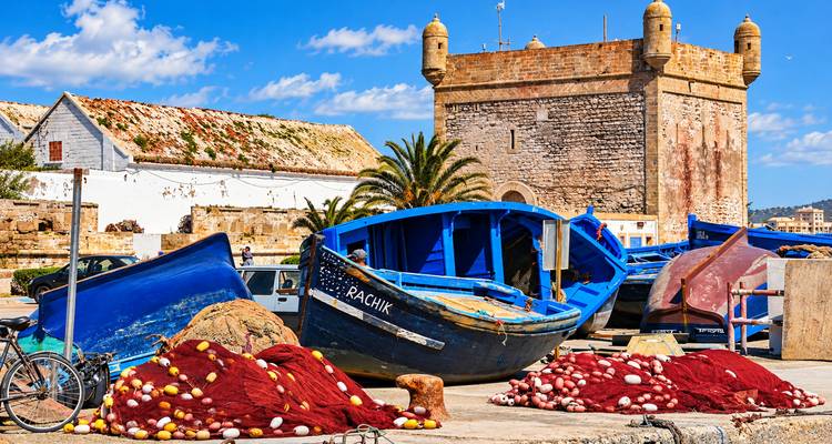 Botes de pesca, redes y una torre de fortaleza de piedra en un día soleado en un puerto costero.