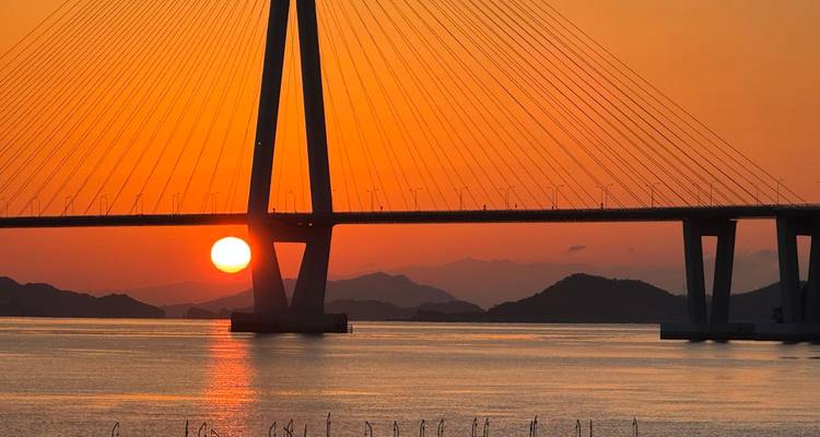 Ein feuerrot-orangener Sonnenuntergang sinkt unter einer Schrägseilbrücke über ruhigen Inselgewässern ab.