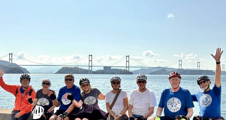 Lächelnde Radfahrergruppe posiert auf einer Ufermauer mit mehreren Inselbrücken im Hintergrund.