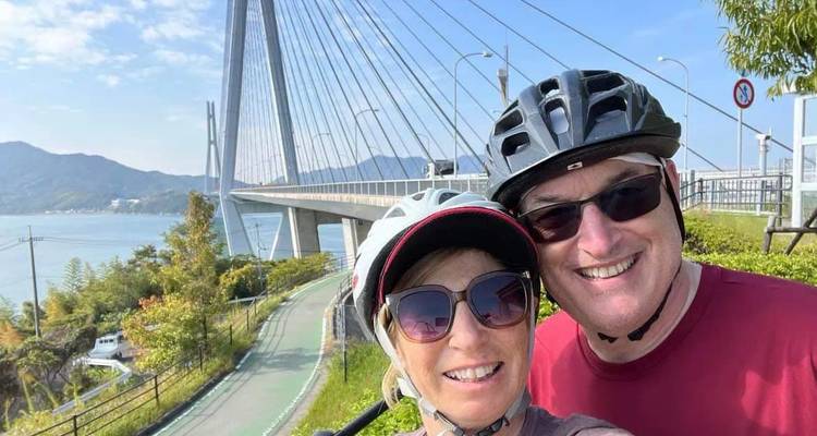 Ein lachendes Paar in Helmen macht ein Selfie in der Nähe einer eleganten Schrägseilbrücke.