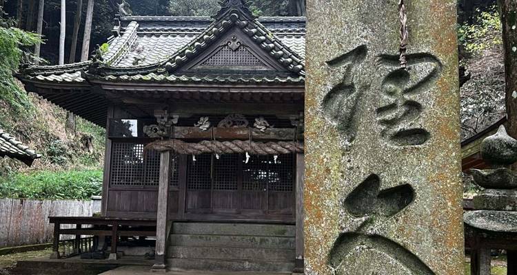 Verwitterte Steinsäule mit Kanji steht vor einem rustikalen Holzbergschrein.