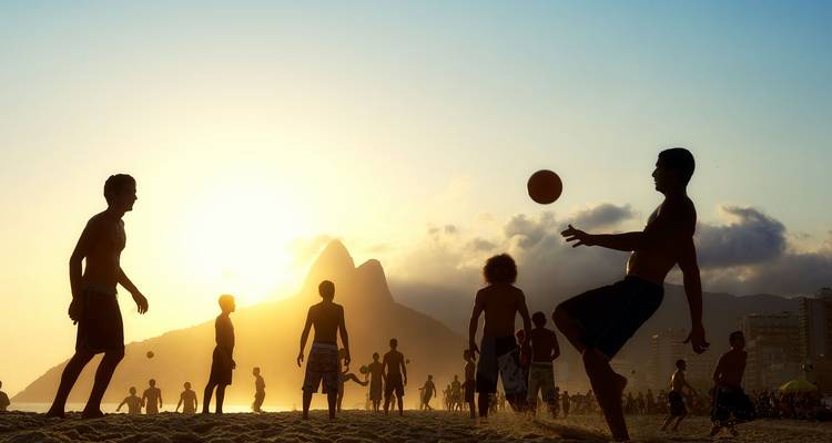 Des silhouettes de personnes jouent au football sur la plage d'Ipanema au coucher du soleil avec les pics des Deux Frères en arrière-plan.