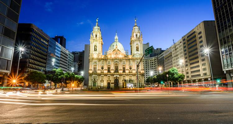 Vue nocturne de l'église illuminée de Candelária de Rio encadrée par des tours de bureaux modernes.