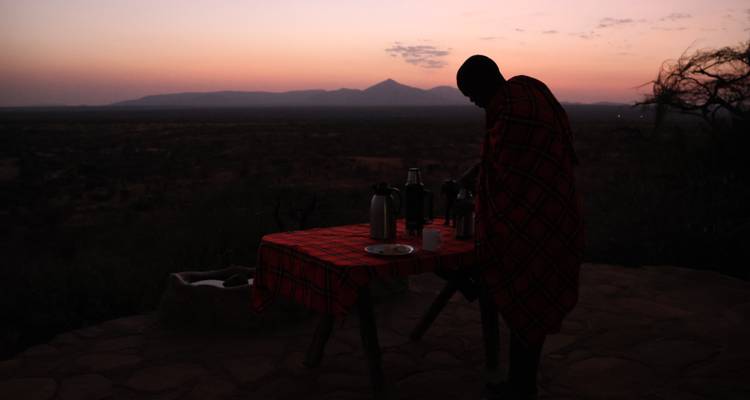 Silhouette of Maasai host preparing coffee on a cliff-edge table at sunrise over savanna.