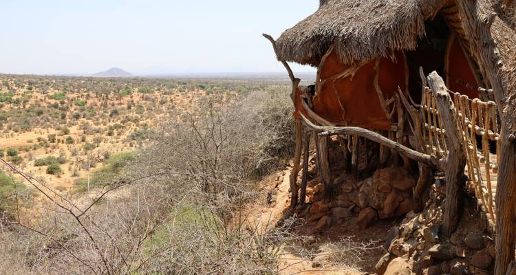 Thatched safari hut perched above arid savanna with distant hills on a clear day.