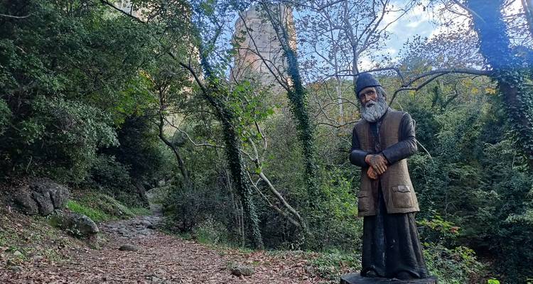 Statue en grandeur nature d'un moine orthodoxe à côté d'un sentier forestier avec les rochers de Météores faiblement visibles.