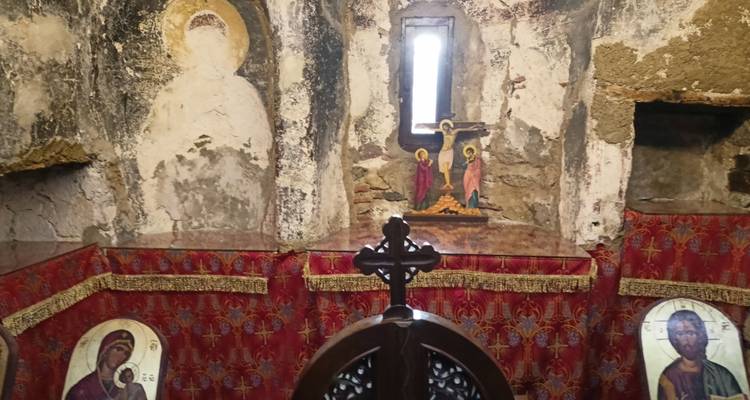 Intérieur faiblement éclairé d'une petite chapelle aux murs rustiques, une tapisserie rouge et une croix en bois.