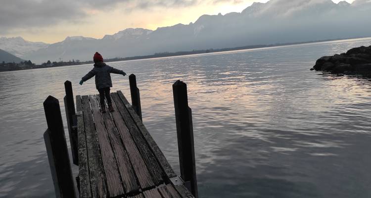 Un enfant se tient en équilibre, les bras tendus, sur une jetée en bois au-dessus d'eaux calmes d'un lac avec des montagnes enneigées à l'horizon.