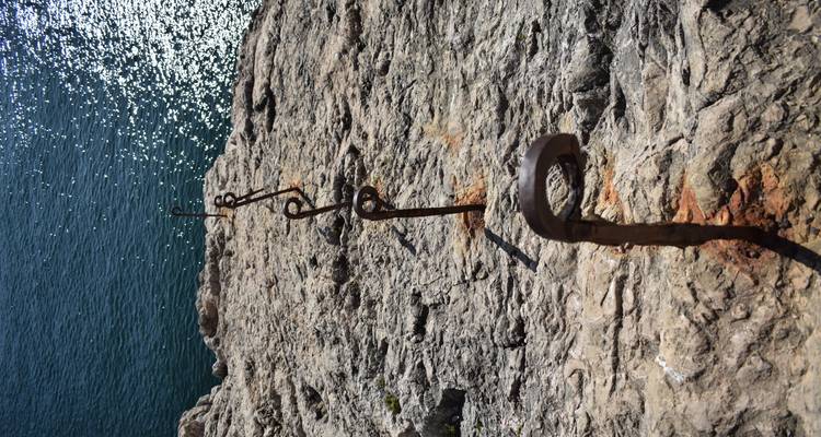 Line of old metal anchor points along a sun-lit cliff edge above sparkling sea