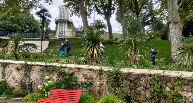 Urbaner Park an einem Hang mit exotischen Pflanzen, spielenden Kindern auf Tretrollern und einem kleinen Aussichtsturm oben.