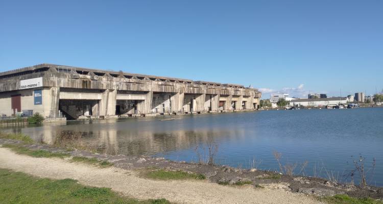 Konkreter U-Boot-Stützpunkt spiegelt sich in ruhigen Becken-Gewässern unter klarem blauen Himmel.