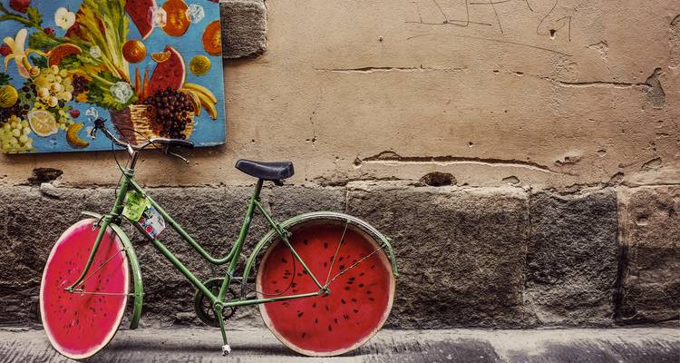 Quirky green bicycle with watermelon-slice wheels leaning against an old stone wall adorned with a colorful fruit painting.
