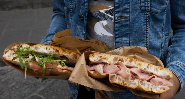 Person holding two stuffed focaccia sandwiches wrapped in brown paper on a city street.