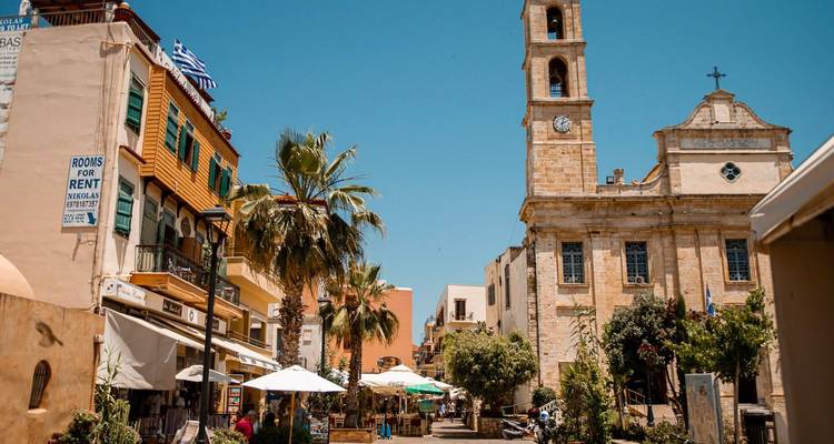 Sonnige Fußgängerzone mit Palmen, Cafes und einer Steinkirche mit Glockenturm in einer historischen griechischen Stadt.